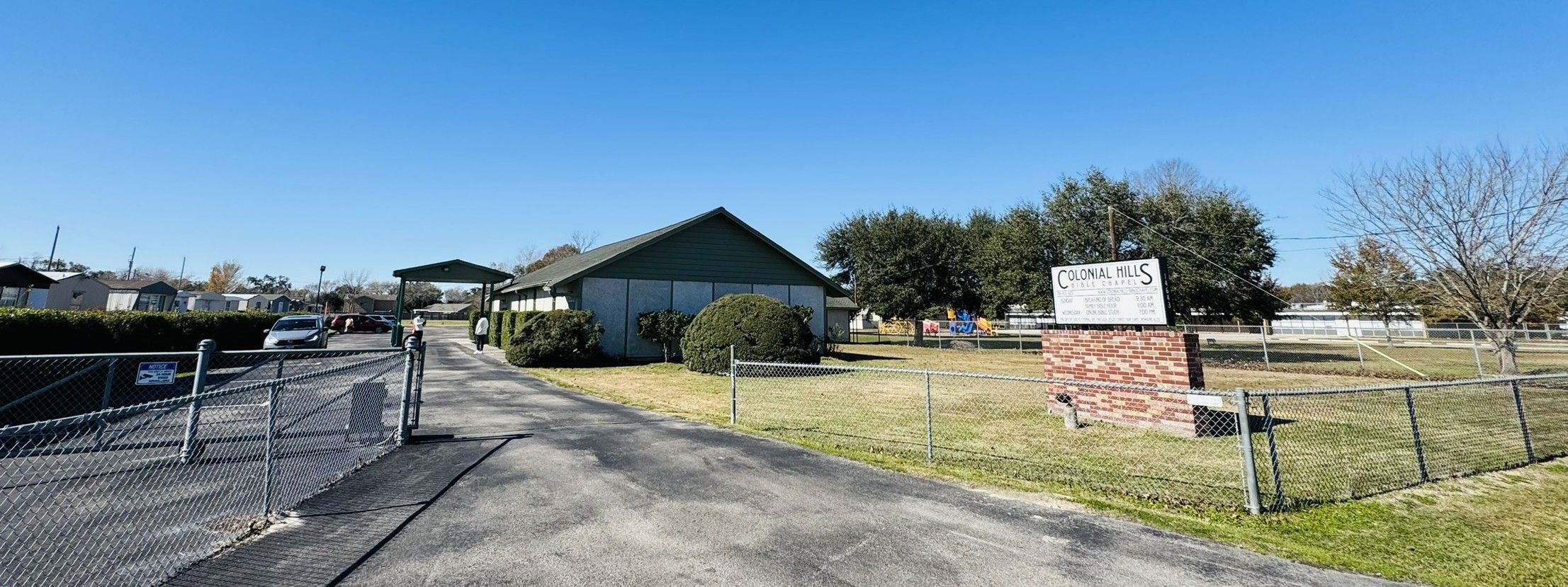 Colonial Hills Bible Chapel seen from its driveway, with the sign and the lawn under a clear blue sky.