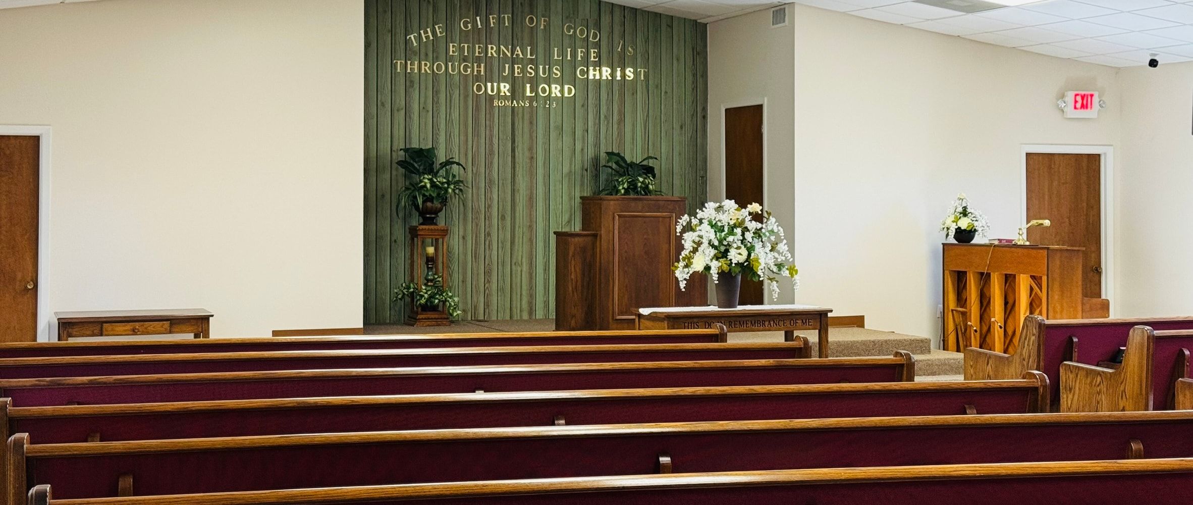 The meeting room of Colonial Hills Bible Chapel: rows of wooden pews facing a green plank wall lettered in gold, “The gift of God is eternal life through Jesus Christ our Lord”.